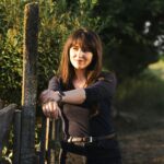 A young woman leans on a fence in the countryside during a beautiful sunset, enjoying the peaceful atmosphere.
